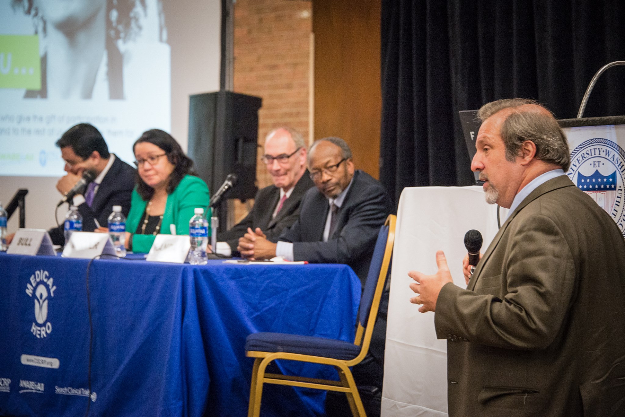 Dr. Mellman holding a microphone speaking at an event in front of four panelists seated on stage.